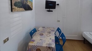 A small, clean room with a dining table, two chairs, a flat-screen TV mounted on the wall, and a door. - VILLA ROSA 2 - Antico Borgo Scopello NEAR THE SEA (Scopello)