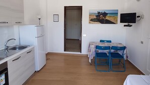A compact kitchen with white cabinets, a sink, and a refrigerator. There is a dining area with a table and chairs, and a painting on the wall. - VILLA ROSA 2 - Antico Borgo Scopello NEAR THE SEA (Scopello)