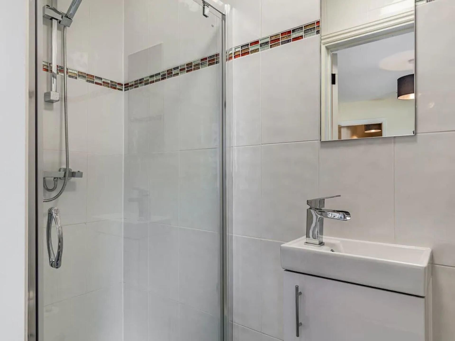 Casa de banho moderna com cabine de duche em vidro, lavatório branco com torneira cromada e painel de azulejos com uma faixa colorida.