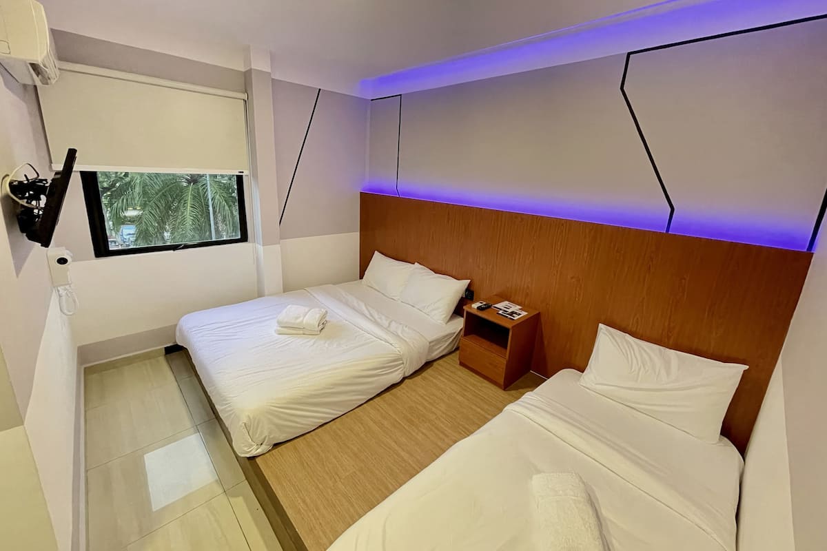Ein Hotelzimmer mit zwei Betten, einem Holzkopfende, einem Nachttisch, einem Fenster mit Blick auf Palmen und einem an der Wand befestigten Fernseher.
