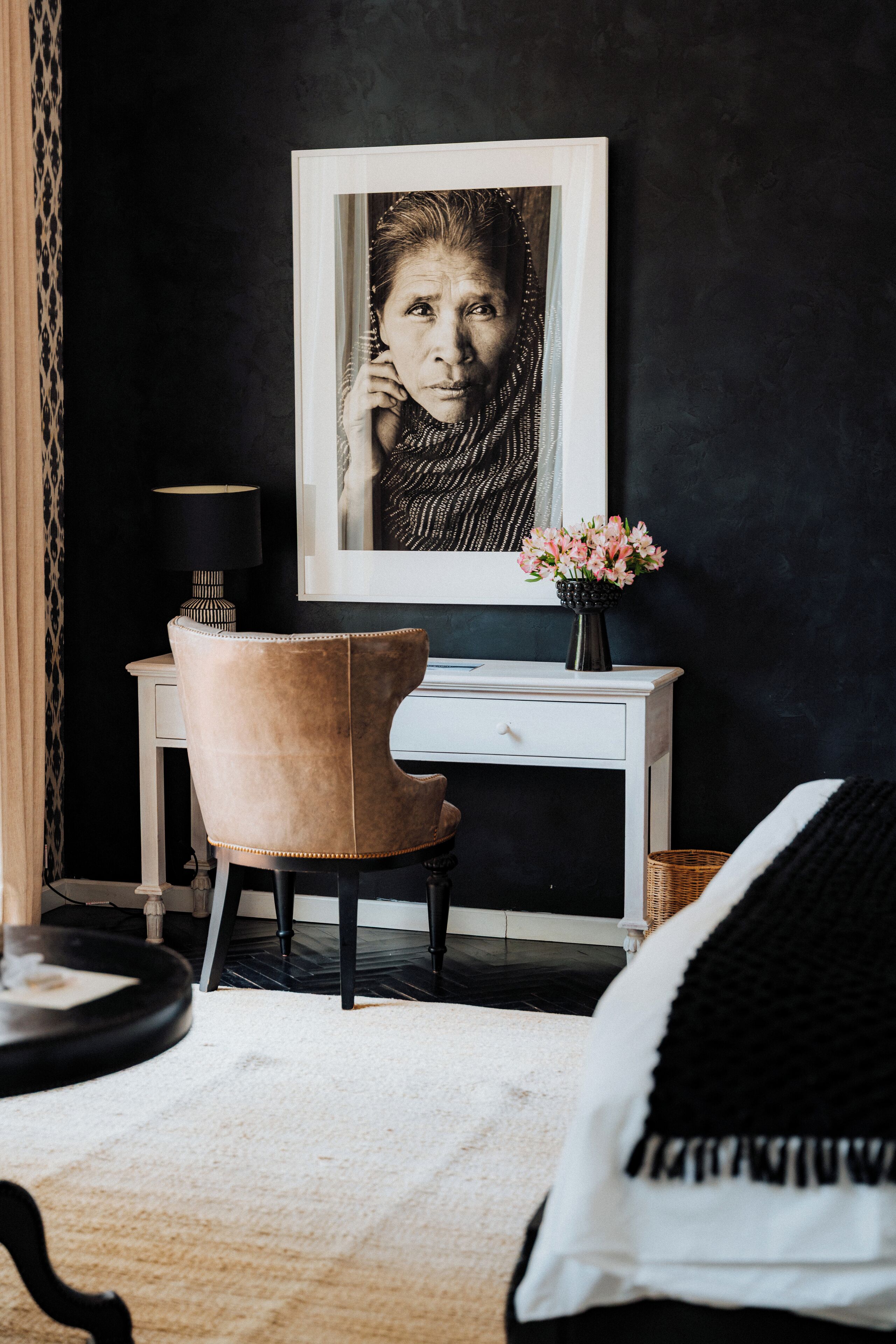 Una habitación con escritorio, silla, un cuadro en la pared, un florero con flores y una lámpara.