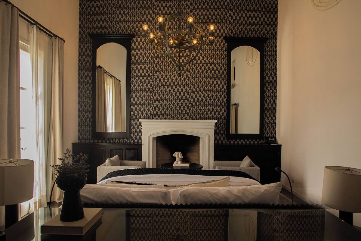 Quarto com cama grande, lareira, duas mesinhas de cabeceira, um candelabro e um espelho amplo.