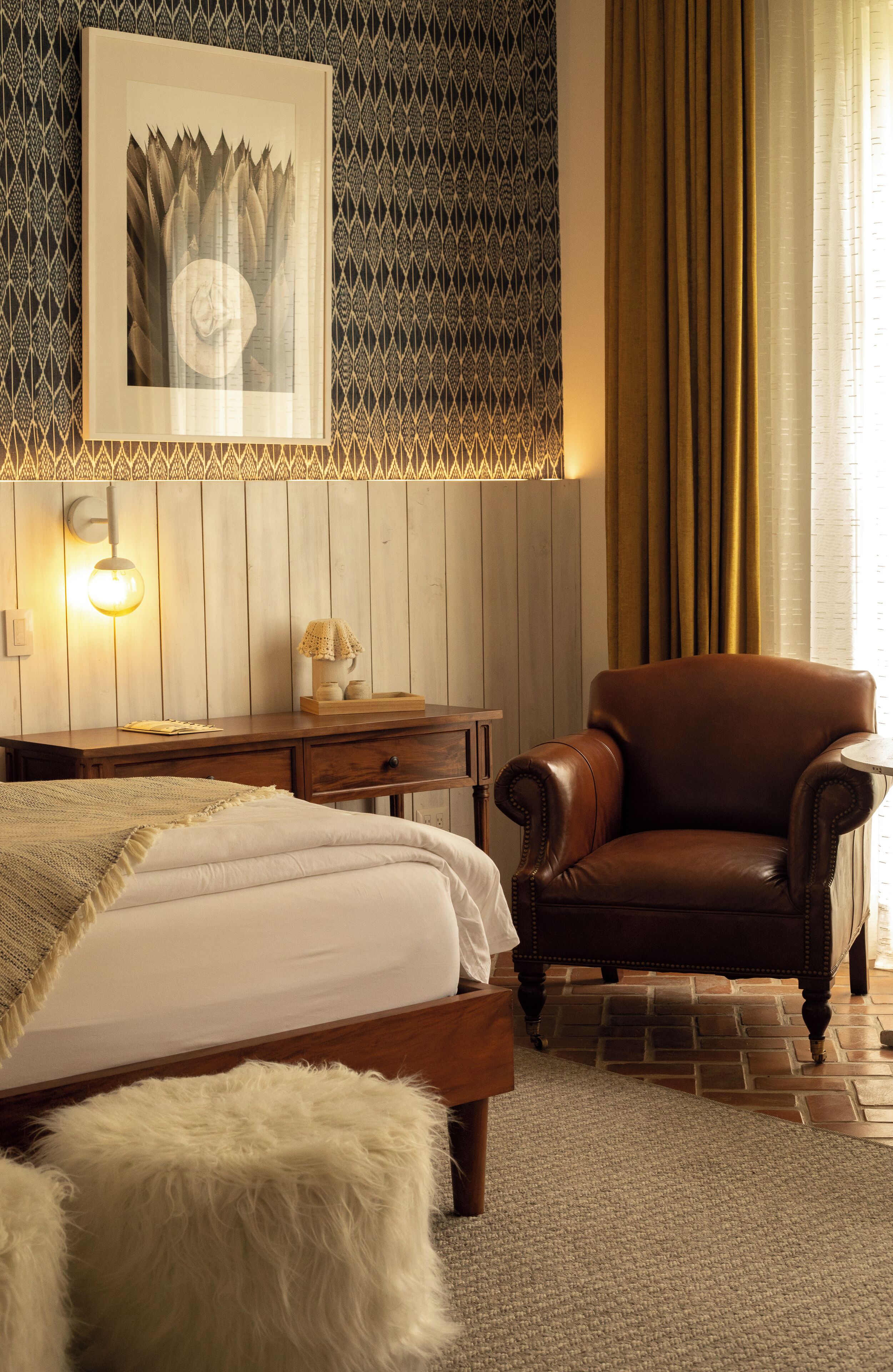 Un dormitorio con cama, un mesita de noche de madera, un sillón de cuero marrón y un papel tapiz estampado.