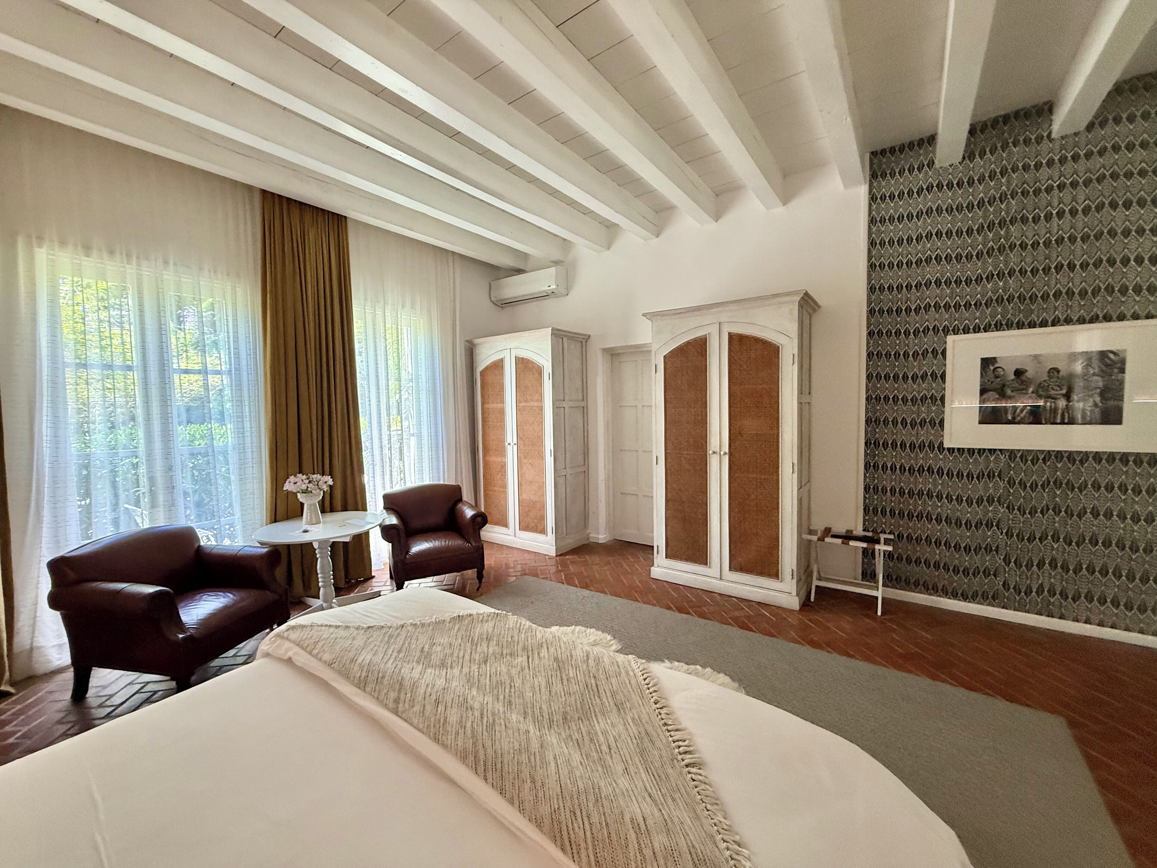 Une chambre d’hôtel avec un grand lit, deux fauteuils, une petite table ornée de fleurs, une armoire et un mur à motifs.