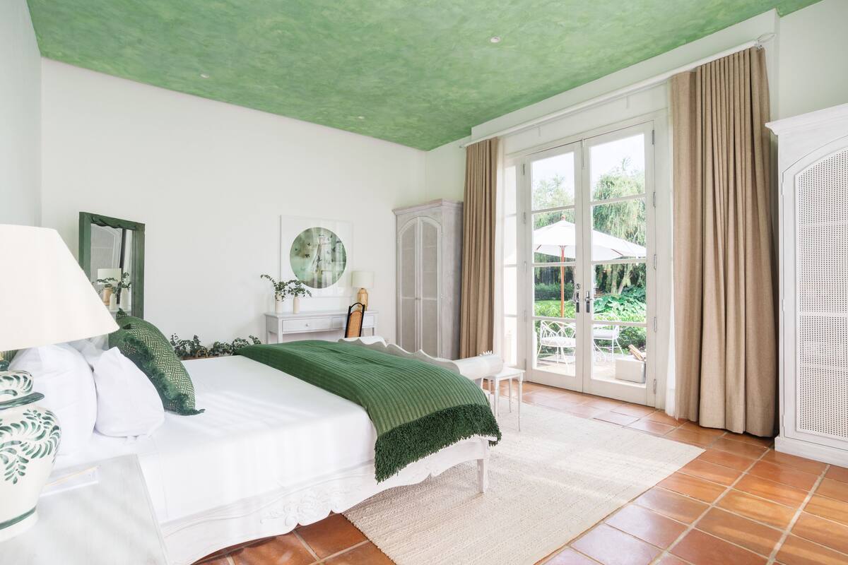 Quarto com cama, mesa de cabeceira, cômoda, espelho e janela com cortinas.