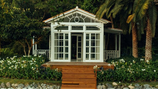 Una casa flotante con terraza de madera y exterior blanco, rodeada de abundante vegetación y una masa de agua tranquila.