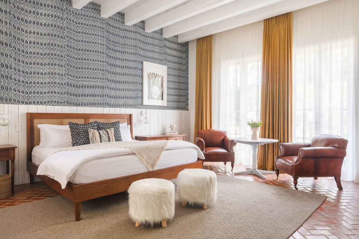 Quarto com uma cama grande, dois pufes brancos e felpudos, uma poltrona de couro marrom, uma mesa redonda pequena e um papel de parede estampado.