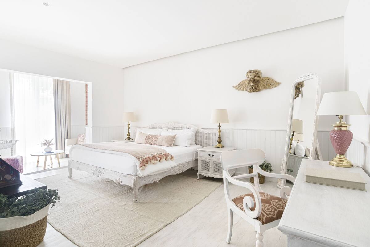 Quarto branco com cama, mesas de cabeceira, uma cadeira, escrivaninha e espelho.