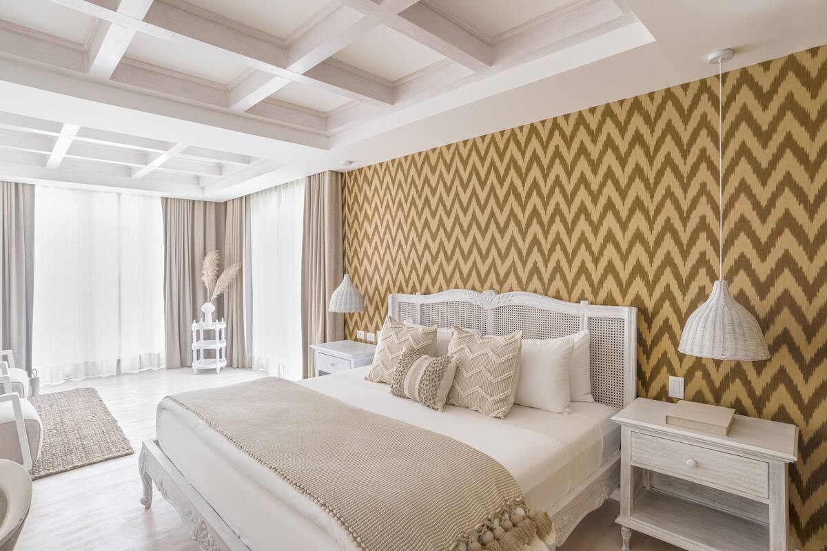 Um quarto espaçoso com uma cama grande, mesas de cabeceira brancas, uma luminária branca e uma parede com padrão em chevron.