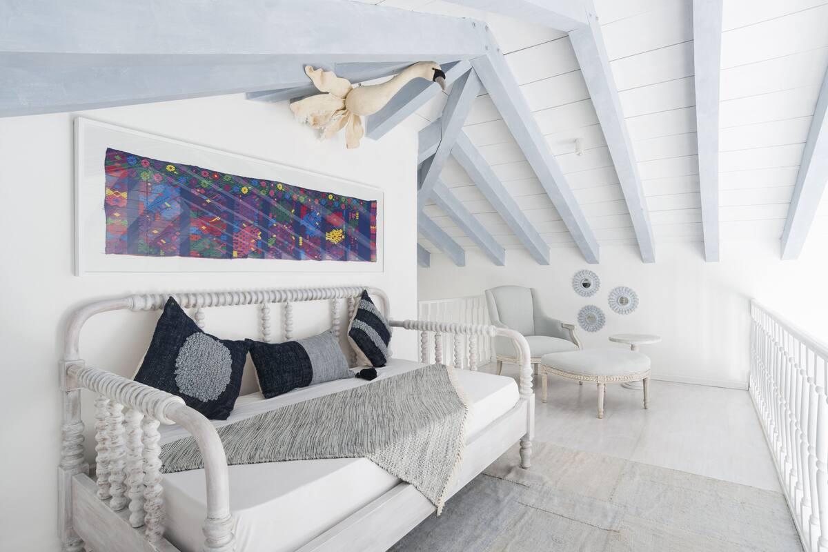 Um sofá branco e ornamentado com manta e almofadas cinzas, uma pintura abstrata emoldurada e uma cadeira branca num quarto claro e branco com teto inclinado.