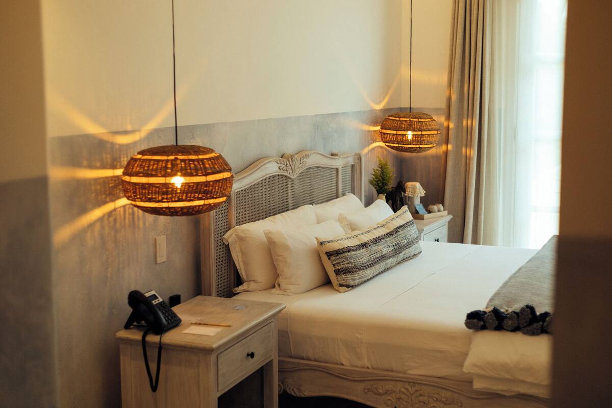 Uma cama bem arrumada com lençóis brancos, uma mesa de cabeceira com abajur e telefone.