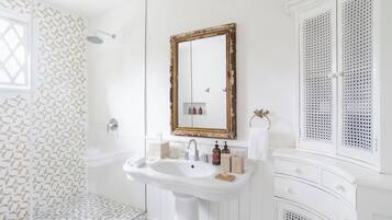 Un baño con lavamanos blanco, espejo y ducha. Hay artículos de aseo en el lavamanos y un perchero en la pared.