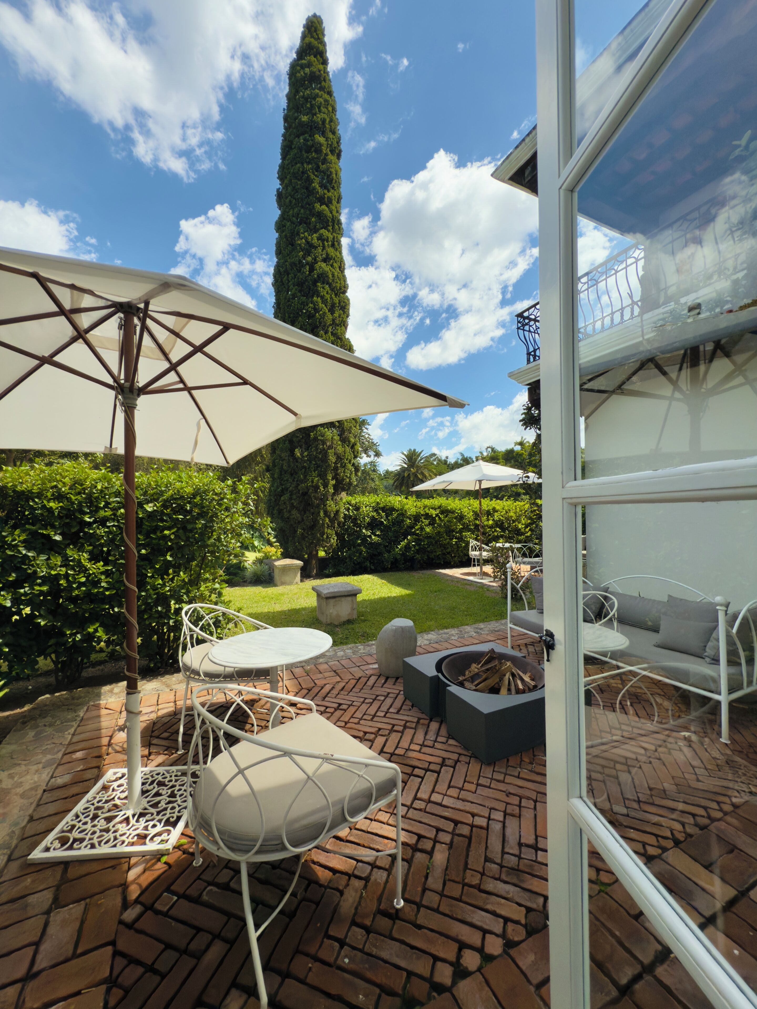 An outdoor patio with a brick floor, white metal furniture, and a large umbrella.