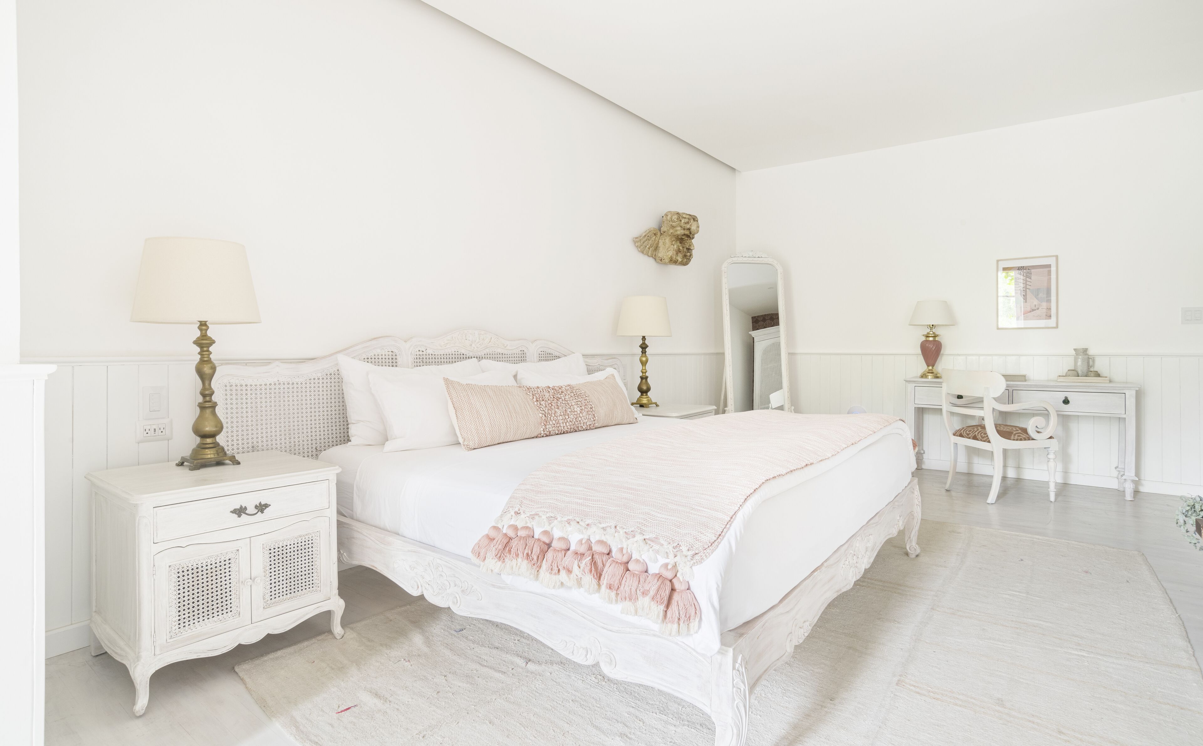 A white bedroom with a large bed, two bedside tables with lamps, a dresser, a mirror, and a chair.