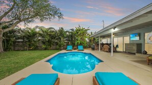 Ein Garten mit Swimmingpool, zwei Liegestühlen und einer überdachten Terrasse mit Fernseher.