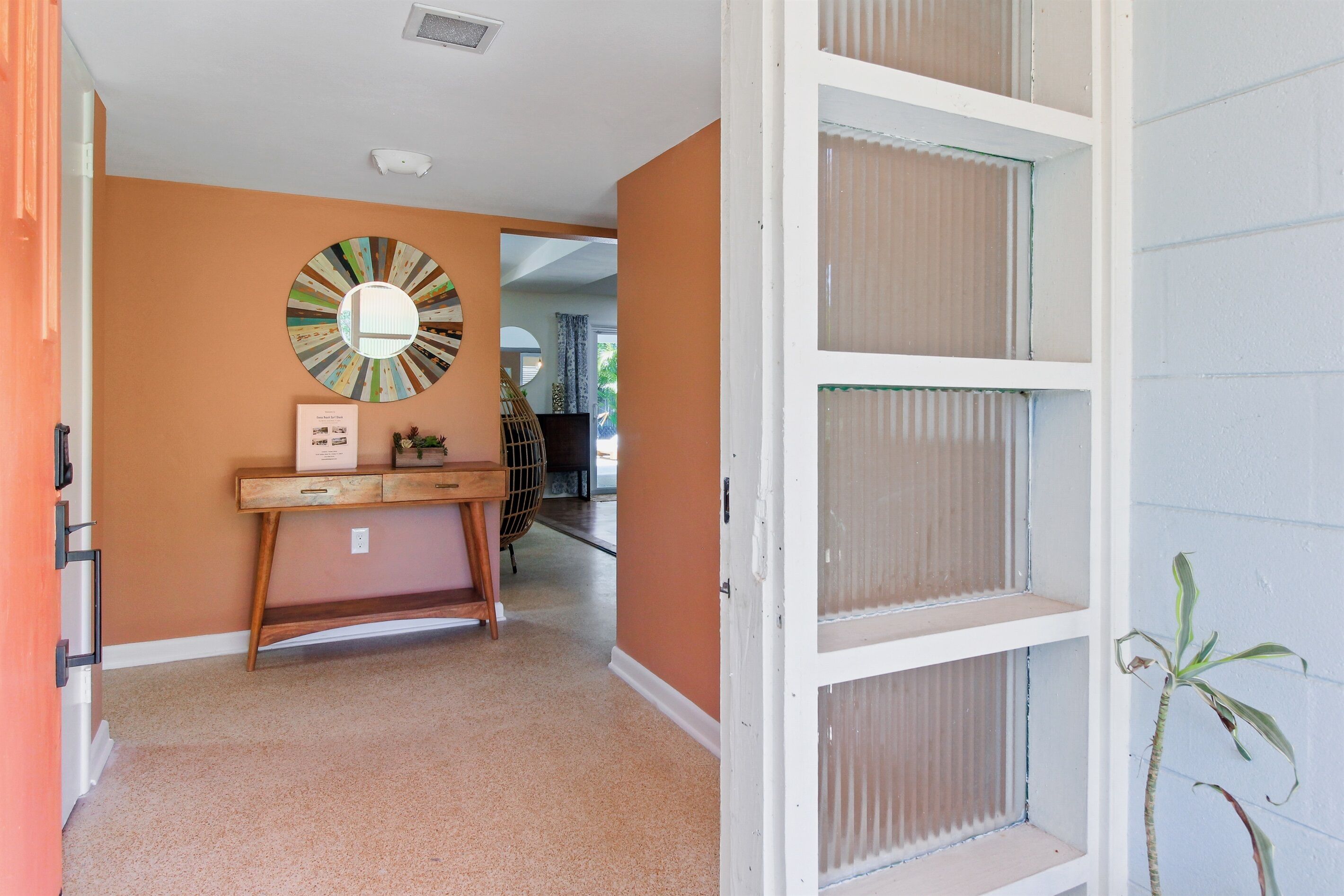A hallway with a wooden console table, a mirror with a colorful frame, and a door with a plant beside it.