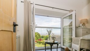 A room with a sliding glass door leading to a balcony with a view of the sea. There is a small table with a vase and a bottle on it. - Sandacre Cottage (Ulverston)