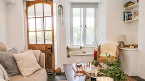 A cozy living room with a wooden door, a window with a view, a sofa, a coffee table with a candle, a bottle of wine, and a plant. - Sandacre Cottage (Ulverston)