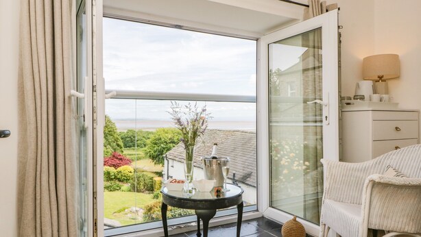 A room with a glass door leading to a garden, a small round table with a vase and a bottle, and a white chair. - Sandacre Cottage (Ulverston)