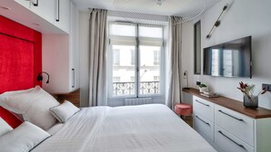 A modern bedroom with a bed, white curtains, a window with a view, a TV, and a bedside table with a plant.