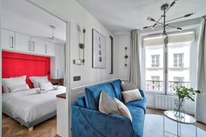Une chambre d’hôtel moderne, équipée d’un lit, d’un canapé bleu, d’une petite table et d’une grande fenêtre donnant sur des immeubles.