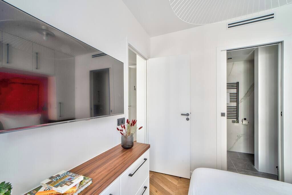 A modern bedroom with a large mirror, a wooden bedside table, a white bed, and a door leading to a bathroom.