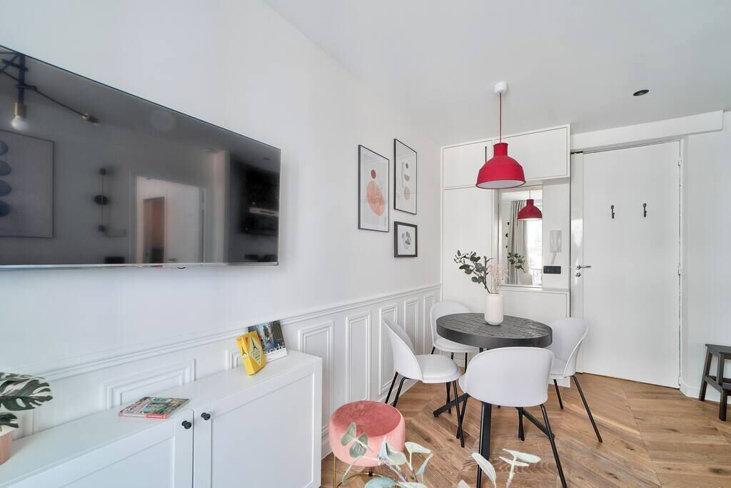 Uma sala de estar moderna com televisão de tela plana, móveis brancos, uma mesa de jantar redonda com cadeiras e uma luminária pendente vermelha.