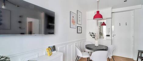 Uma sala de estar moderna com televisão de tela plana, móveis brancos, uma mesa de jantar redonda com cadeiras e uma luminária pendente vermelha.