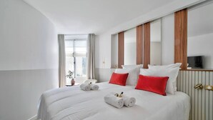 Quarto de hotel com uma cama grande, roupa de cama branca, almofadas vermelhas e uma TV na parede.