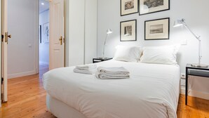 Ein ordentlich bezogenes Bett mit weißen Leinen und Handtüchern, ein Nachttisch mit Lampe und gerahmte Bilder an der Wand.