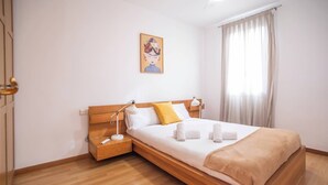 A hotel room with a wooden bed, a bedside table, a nightstand, a lamp, a painting on the wall, and a window with curtains.