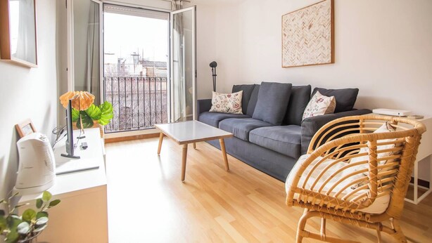 A modern living room with a grey sofa, a wooden coffee table, a wicker chair, and a large window with a view of buildings.