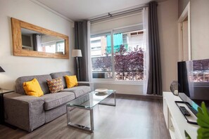 Un salon moderne avec un canapé gris, une table basse en verre, un téléviseur et une grande fenêtre donnant sur des immeubles et des arbres.