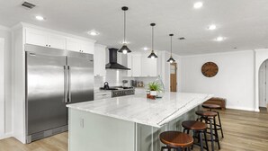 Une cuisine moderne dotée d’un grand îlot central, d’appareils électroménagers en acier inoxydable, de luminaires suspendus et d’une horloge en bois accrochée au mur.