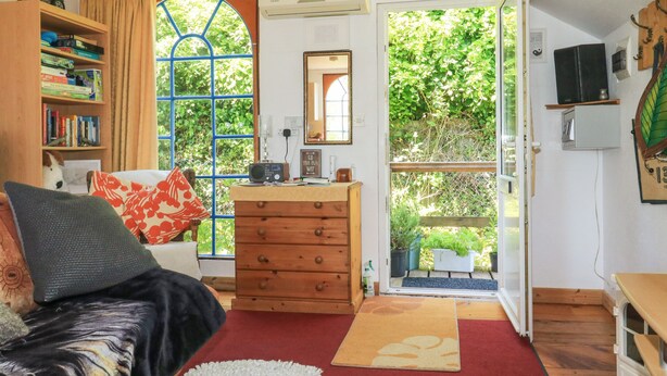 A cozy living room with a bookshelf, a wooden dresser, a red and white rug, and a view of greenery outside. - Detox Dunchideock (Exeter)