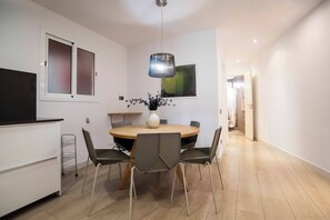 A modern dining area with a round table, six chairs, a vase with flowers, a hanging light fixture, and a TV on the wall.