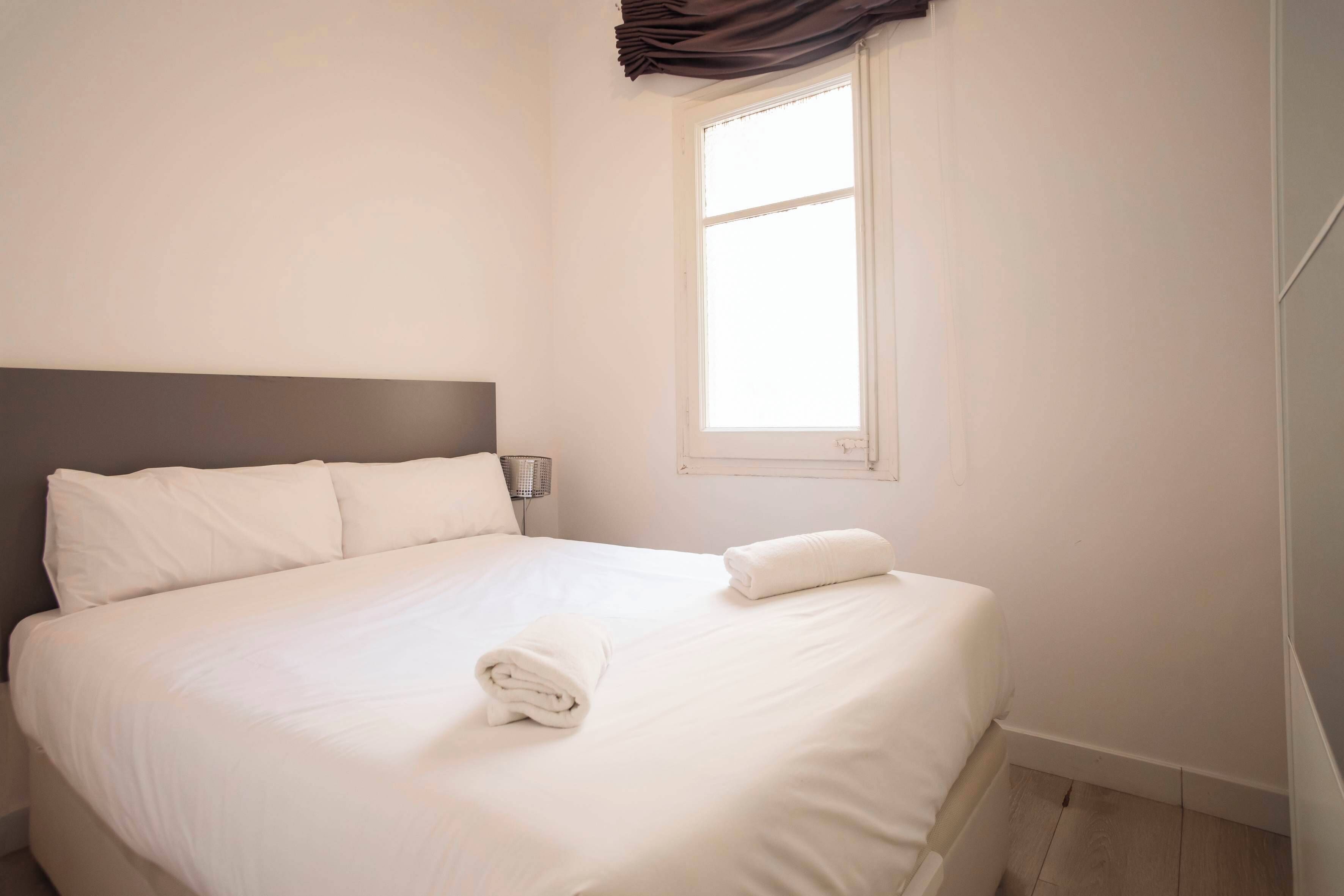 Una cama bien hecha con sábanas blancas y una toalla doblada, una lámpara de noche y una ventana con cortinas.