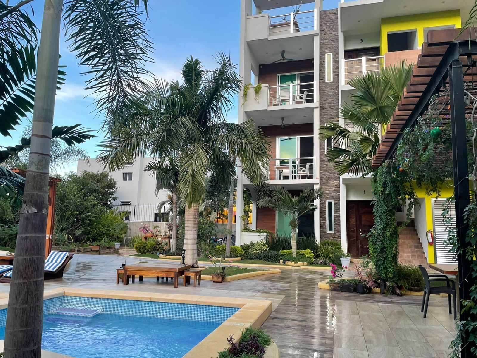 Un edificio moderno de varios pisos con piscina, mobiliario exterior y abundante vegetación.