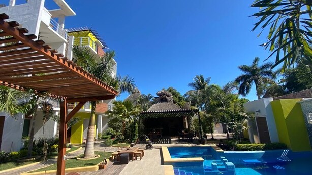 A resort pool area with a wooden pergola, lounge chairs, and a thatched-roof structure.