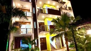 A multi-story building with balconies, a well-lit entrance, and a landscaped garden with palm trees.