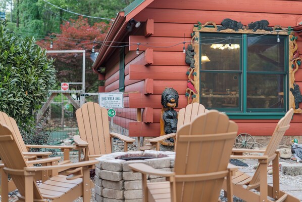 A wooden cabin with a dog walk area sign, outdoor seating with Adirondack chairs, and a stone fire pit.