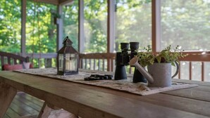 Una mesa de madera con binoculares, un farol y una planta en maceta en un porche con vistas a árboles.