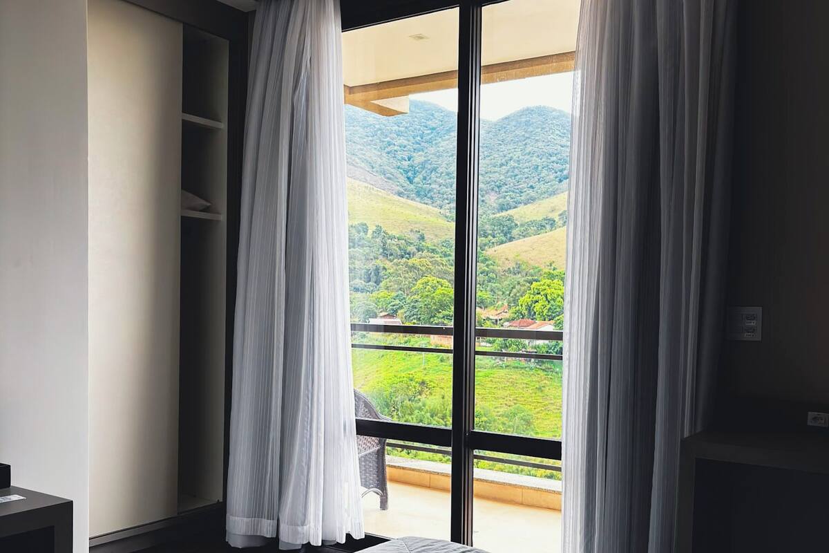 A bedroom with a large bed, a sliding glass door leading to a balcony, and a view of green hills.