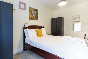 A neatly made bed with white linens and a yellow accent pillow, a dark wooden wardrobe, a framed artwork on the wall, and a window with blinds. - Central 3BD Home Short walk to Edinburgh Old Town (Edinburgh)