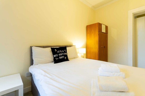 A neatly made bed with white linens, a black pillow with text, a wooden wardrobe, and a white bedside table.