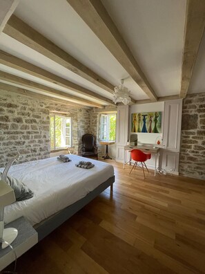 Ein Schlafzimmer mit einem großen Bett, einem Schreibtisch und einem Stuhl. Eine Steinsäule und eine Holzbalkendecke sind ebenfalls vorhanden.
