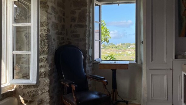 Une chambre avec un mur en pierre, un fauteuil en cuir noir, un lit recouvert d’une couverture à rayures et une fenêtre donnant sur un paysage verdoyant.