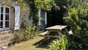 Un bâtiment en pierre avec des volets blancs, une table de pique-nique en bois et une végétation luxuriante.