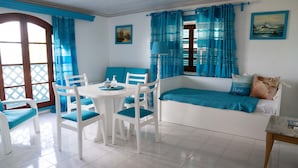 A room with a white table and chairs, a blue cushioned bench, and a view of the sea through blue curtains.