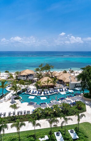 A resort with a central pool surrounded by lounge chairs and umbrellas, a beach area with thatched-roof structures, and a view of the ocean.